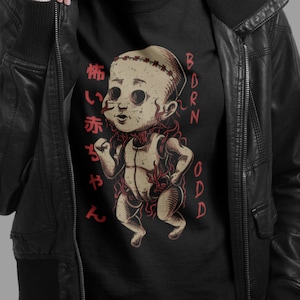 May include: Black t-shirt featuring a graphic of a stitched-up baby with red vines growing from its body. The text "BORN ODD" is on the right side of the graphic and Japanese text is on the left side.