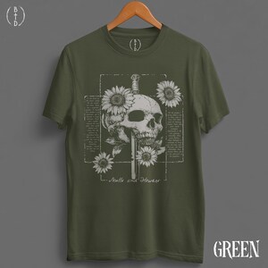 May include: Olive green t-shirt featuring a detailed graphic of a skull pierced by a sword, surrounded by sunflowers. The design includes the text "Skulls and Flowers" in a vintage style. The shirt is displayed on a wooden hanger.