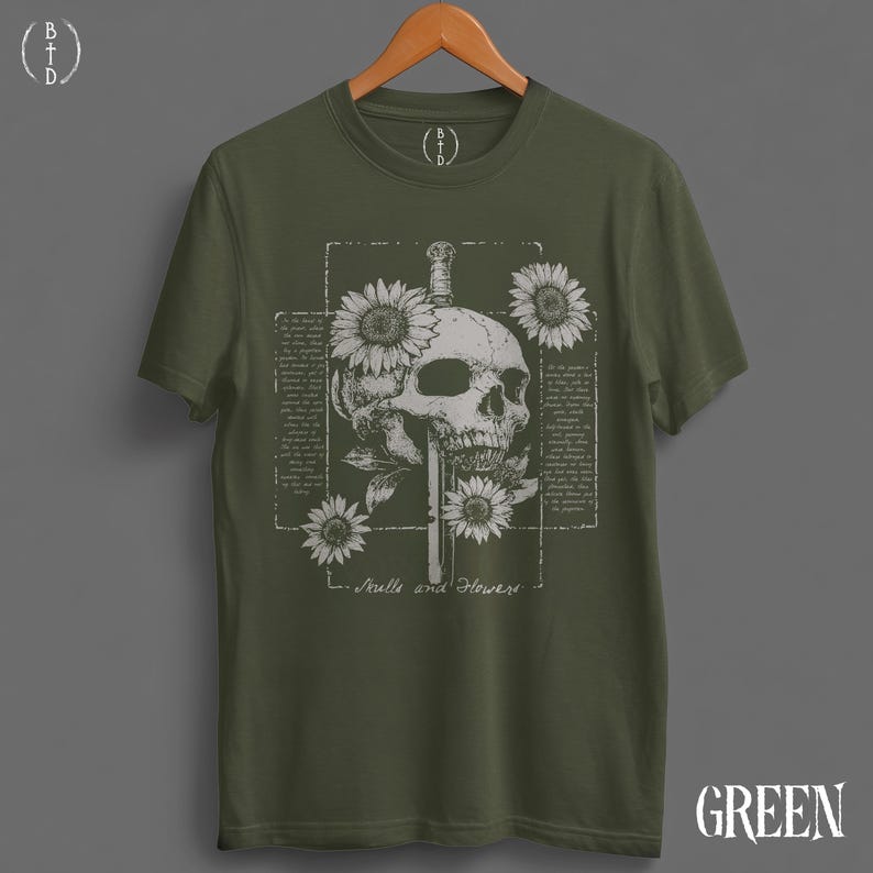 May include: Olive green t-shirt featuring a detailed graphic of a skull pierced by a sword, surrounded by sunflowers. The design includes the text "Skulls and Flowers" in a vintage style. The shirt is displayed on a wooden hanger.