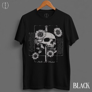 May include: Black t-shirt featuring a detailed skull pierced by a sword, surrounded by sunflowers. The design includes the text "Skulls and Flowers" and a rectangular frame with text. The word "BLACK" is printed at the bottom right.