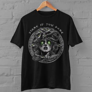 Op de afbeelding: Zwart T-shirt met een grafische print van Medusa, een mythische Griekse figuur met slangenhaar. De tekst "Stare If You Dare" is in een cirkel rond de afbeelding gedrukt.
