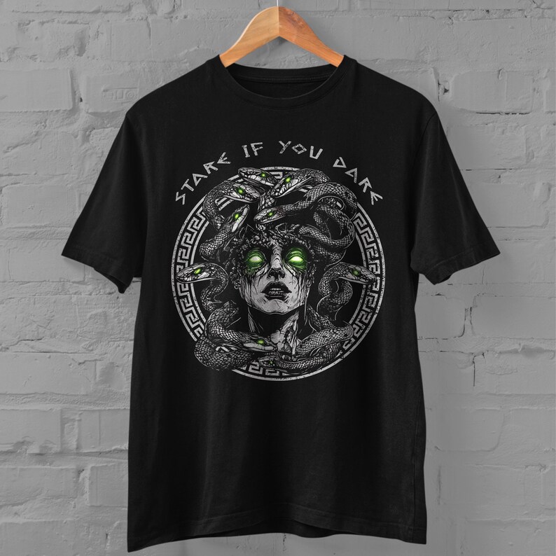 Op de afbeelding: Zwart T-shirt met een grafische print van Medusa, een mythische Griekse figuur met slangenhaar. De tekst "Stare If You Dare" is in een cirkel rond de afbeelding gedrukt.