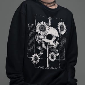 May include: A black long-sleeve sweatshirt featuring a white graphic print of a human skull pierced by a sword, surrounded by sunflowers and blocks of illegible cursive text. The bottom of the design includes the text Skulls and Flowers.