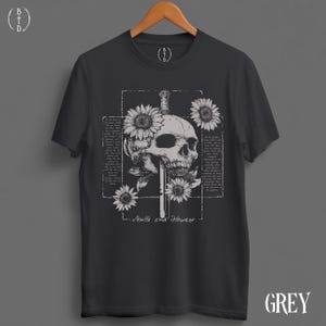 May include: Dark grey t-shirt featuring a skull pierced by a sword, surrounded by sunflowers. The design includes text within a rectangular frame and the words "Skulls and Flowers" below the graphic. The word "GREY" is printed on the bottom right.