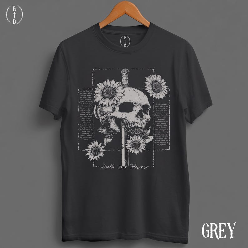 May include: Dark grey t-shirt featuring a skull pierced by a sword, surrounded by sunflowers. The design includes text within a rectangular frame and the words "Skulls and Flowers" below the graphic. The word "GREY" is printed on the bottom right.