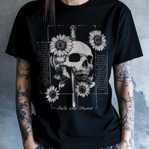 May include: Black t-shirt featuring a detailed skull pierced by a sword, surrounded by sunflowers. The design includes text within a rectangular frame, with the words "Skulls and Flowers" at the bottom. The overall aesthetic is gothic.