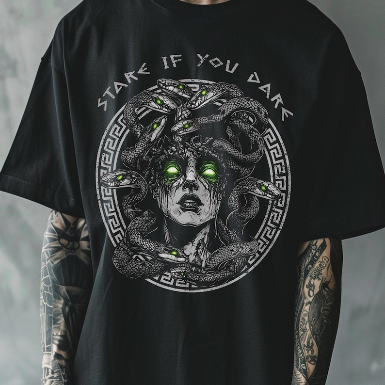 Op de afbeelding: Zwart T-shirt met een grafische print van Medusa, een mythische Griekse figuur met slangenhaar. De tekst "Stare If You Dare" is in een cirkel rond de afbeelding gedrukt.