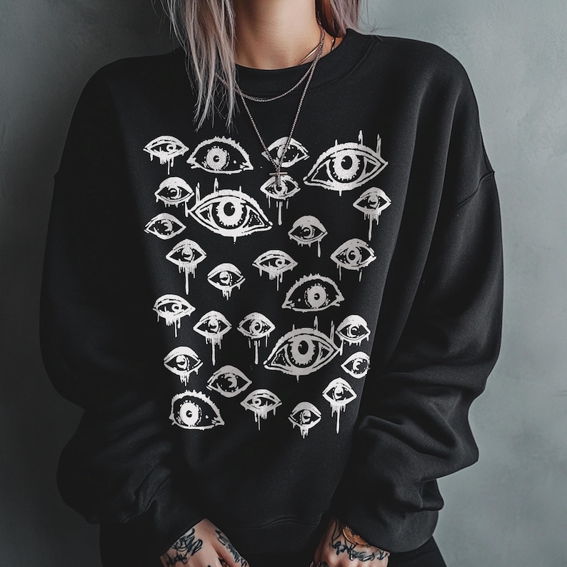 May include: A black long-sleeve sweatshirt featuring a white graphic print of numerous eyes of varying sizes. The eyes are depicted in a sketch-like style with some appearing to drip or bleed downward. The sweatshirt has a crew neckline.