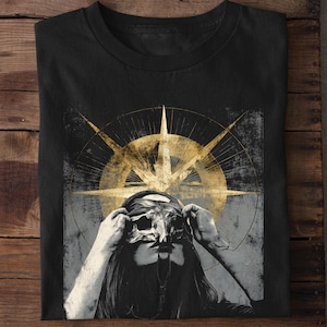Puede incluir: Camiseta negra con un gr&aacute;fico desgastado de una persona sosteniendo una m&aacute;scara, con un dise&ntilde;o de br&uacute;jula dorada en el fondo. El dise&ntilde;o tiene una est&eacute;tica vintage y grunge. La camiseta est&aacute; hecha de un material suave.