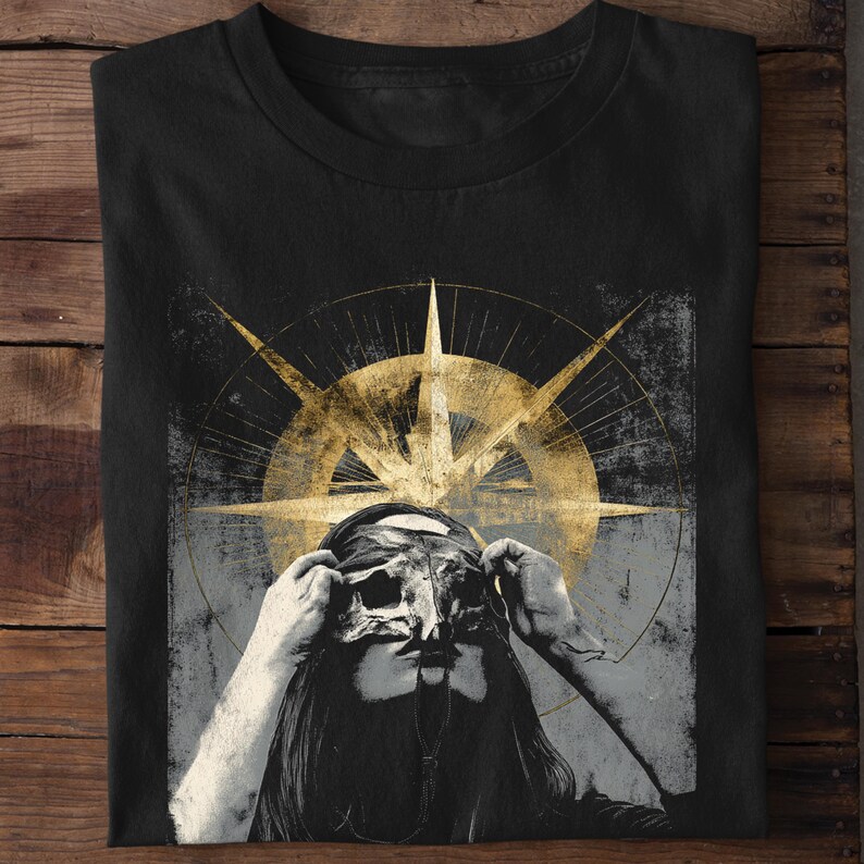 Puede incluir: Camiseta negra con un gr&aacute;fico desgastado de una persona sosteniendo una m&aacute;scara, con un dise&ntilde;o de br&uacute;jula dorada en el fondo. El dise&ntilde;o tiene una est&eacute;tica vintage y grunge. La camiseta est&aacute; hecha de un material suave.