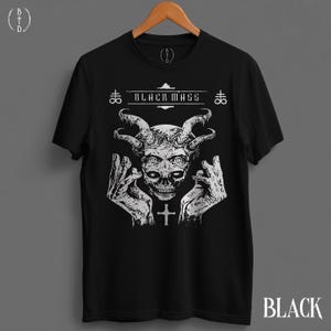 May include: A black short-sleeve t-shirt featuring a white graphic print of a horned skull with hands positioned on either side. The text "BLACK MASS" is printed above the skull, flanked by two symbols, with a small cross below the skull.
