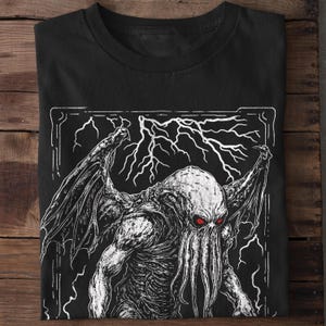 K&ouml;nnte beinhalten: Schwarzes T-Shirt mit einem wei&szlig;en Grafikdruck eines tentakelartigen Wesens mit roten Augen. Das Wesen ist von Blitzen umgeben und von einem wei&szlig;en Rahmen eingefasst.