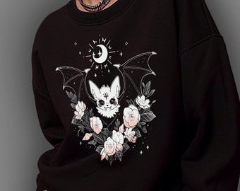 May include: A black crewneck sweatshirt featuring a white graphic print of a bat with large ears, surrounded by blooming flowers and a crescent moon with stars above its head. The design is centered on the chest of the long-sleeved garment.