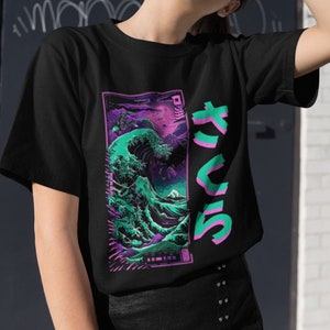May include: A black short-sleeve t-shirt featuring a rectangular graphic of a stylized teal and purple ocean wave with a mountain in the background. To the right of the graphic, large Japanese characters are printed in a bright teal color.