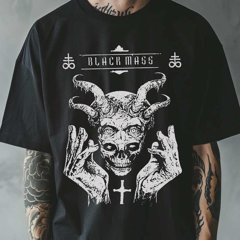 May include: A black short-sleeve t-shirt featuring a white graphic print of a horned skull with hands raised on either side. The text "BLACK MASS" is printed above the skull, flanked by two symbols, with a cross icon below the skull.