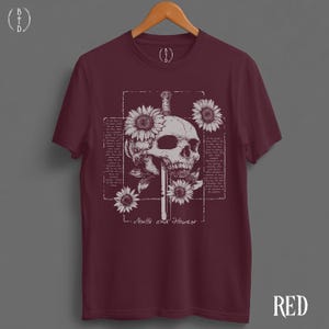 May include: Burgundy t-shirt featuring a skull design with a sword, sunflowers, and text that reads "Skulls and Flowers." The graphic is in white, set against a dark red background. The shirt is displayed on a wooden hanger.