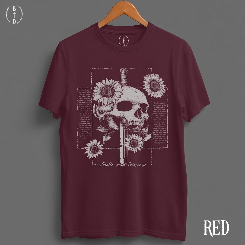 May include: Burgundy t-shirt featuring a skull design with a sword, sunflowers, and text that reads "Skulls and Flowers." The graphic is in white, set against a dark red background. The shirt is displayed on a wooden hanger.