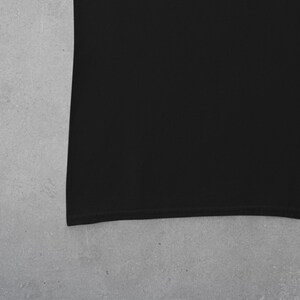 Puede incluir: Una camiseta negra lisa, con la textura de la tela visible. El borde de la camiseta es curvo, lo que sugiere un ajuste relajado. El fondo es gris claro, contrastando con la prenda oscura.