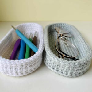 Könnte beinhalten: Zwei handgefertigte Häkelkörbe, einer weiß und einer grau. Der weiße Korb enthält Häkelnadeln in verschiedenen Farben, darunter Blau, Grün und Lila. Der graue Korb enthält eine Brille. Beide Körbe sind oval geformt.