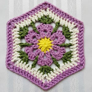 Op de afbeelding: Een handgemaakte gehaakte zeshoek in een bloemenontwerp. Het midden is geel, omgeven door paarse, witte en groene bloemblaadjes. De buitenrand is effen paars, waardoor een decoratief stuk ontstaat.