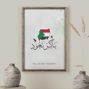 Puede incluir: Impresión artística enmarcada con el mapa de Sudán en los colores de la bandera sudanesa, caligrafía árabe y el texto en inglés «We'll Return Tomorrow». La impresión está en un marco de madera.
