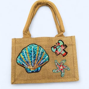 May include: A tan burlap tote bag with two handles and three sequined patches: a seashell, a lobster, and a starfish. The seashell is blue and gold. The lobster is red, pink, and green. The starfish is red and blue.