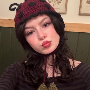 May include: A maroon and black crocheted hat featuring a ladybug-inspired design with black spots, two curled antennae on top, and two dangling olive green leaf-shaped tassels attached to braided cords hanging from the sides.