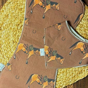 Op de afbeelding: Een bruine baby slabbetje en spuugdoek set. De set heeft een herhaald patroon van een paard met een zadel en hoefijzer. De slab en doek liggen op een geweven, cirkelvormige mat, tegen een houten achtergrond.