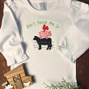 May include: White long-sleeved baby bodysuit with the embroidered phrase "Don't Fence Me In." The design features a black cow, a pink pig, and a red rooster. The bodysuit has a round neckline and ruffled sleeves.