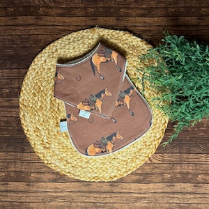 Op de afbeelding: Twee bruine baby slabbetjes met een paard-en-zadel patroon. De slabbetjes liggen op een geweven mat met een houten achtergrond en groen. De slabbetjes hebben een witte rand.