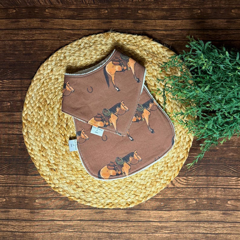 Op de afbeelding: Twee bruine baby slabbetjes met een paard-en-zadel patroon. De slabbetjes liggen op een geweven mat met een houten achtergrond en groen. De slabbetjes hebben een witte rand.