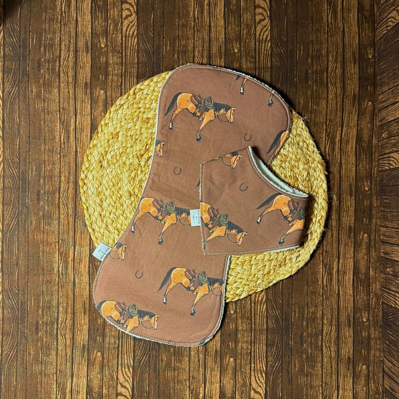 Op de afbeelding: Een bruine baby slabbetje en spuugdoek set. De set heeft een herhaald patroon van een paard met een zadel en hoefijzer. De slab en doek liggen op een geweven, cirkelvormige mat, tegen een houten achtergrond.