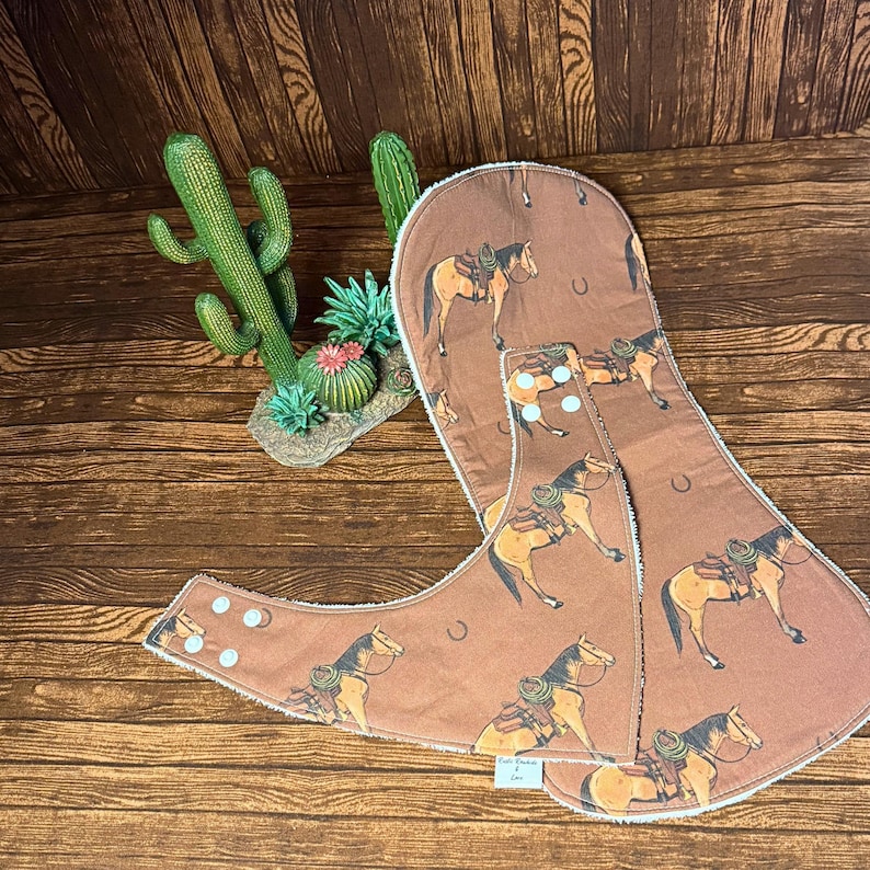 Op de afbeelding: Bruine baby slabbetjes met een herhaald patroon van een paard met zadel. De slabbetjes hebben een witte rand en zijn voorzien van knopen. Een cactus- en vetplantdecoratie staat op de achtergrond.