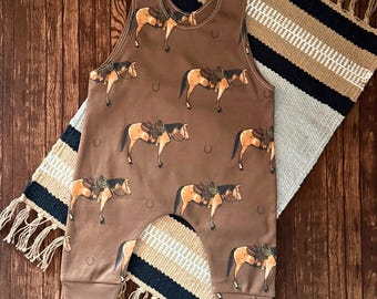 May include: Brown baby romper with a repeating pattern of horses wearing saddles and cowboy hats. The romper has snap closures on the shoulders and a horseshoe design. The romper is on a brown wooden surface with a fringed rug.