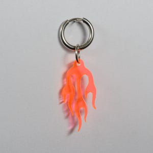 Könnte beinhalten: Ein einzelner silberner Creolen-Ohrring mit einem leuchtend orangefarbenen, flammenförmigen Anhänger. Das Flammendesign ist durchscheinend und wirft einen weichen Schatten. Der Ohrring wird vor einem schlichten weißen Hintergrund gezeigt.