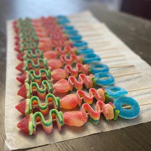May include: A collection of candy skewers arranged in rows on parchment paper. Each skewer features a pointed red and green gummy tip, a rainbow-colored sour ribbon, a round peach-flavored gummy, and a blue sugar-coated gummy ring.