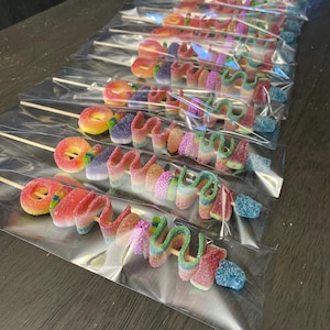 May include: Rows of colorful candy skewers, each individually wrapped in clear plastic. The skewers feature a variety of gummy candies, including rings, worms, and fruit slices, in vibrant rainbow colors. A sweet treat.