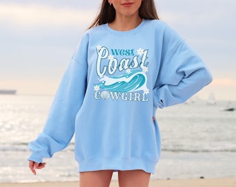 May include: A light blue sweatshirt with a graphic design that says "West Coast Cowgirl" with a wave and seashells. The graphic is in teal and white.