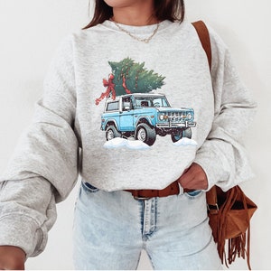 May include: A light gray sweatshirt featuring a blue vintage SUV with a Christmas tree tied to the roof. The tree is green with a red bow. The SUV is parked in a snowy scene.