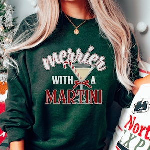 May include: Dark green sweatshirt with the text "Merrier with a Martini" in white and red. The design includes a martini glass with a candy cane and a red bow. The sweatshirt is a festive holiday apparel item.