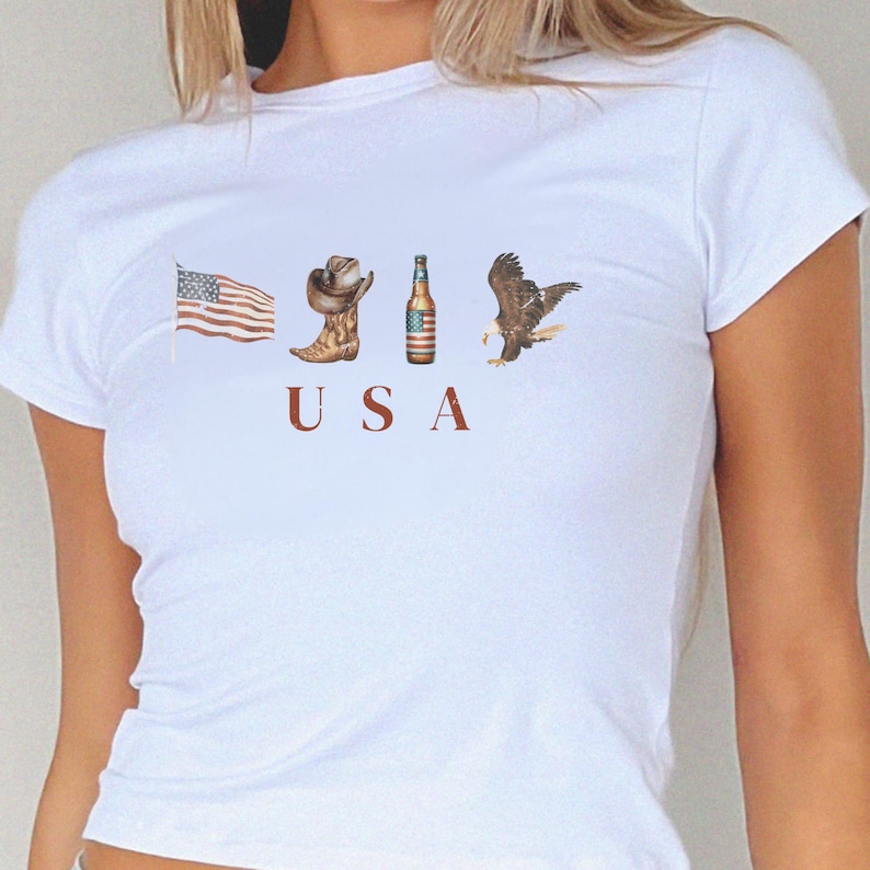 Peut inclure: Un t-shirt blanc &agrave; manches courtes avec une impression graphique repr&eacute;sentant un drapeau am&eacute;ricain, une botte de cow-boy marron, une bouteille de bi&egrave;re avec une &eacute;tiquette drapeau am&eacute;ricain et un pygargue &agrave; t&ecirc;te blanche. Les lettres U S A sont imprim&eacute;es en rouge.