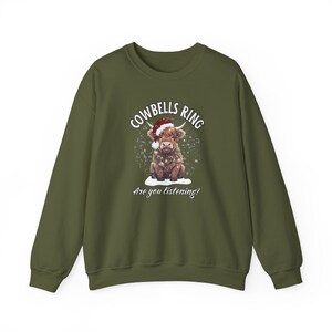 May include: Olive green sweatshirt featuring a festive Highland cow illustration. The cow wears a Santa hat and Christmas lights, with the text "Cowbells Ring, Are you listening?" in white. A cozy, seasonal design.