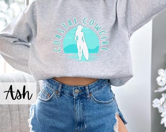 May include: A gray sweatshirt with the text "Coastal Cowgirl" in a turquoise blue circle. The circle features a silhouette of a woman in a cowboy hat standing on a surfboard.