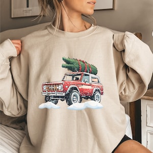 May include: Beige sweatshirt featuring a red vintage SUV with a Christmas tree strapped to the roof. The graphic includes snow and a festive holiday theme. The sweatshirt has long sleeves and a crew neck.