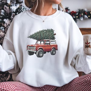 May include: White sweatshirt featuring a red vintage SUV carrying a green Christmas tree on its roof. The graphic design is a festive holiday theme, perfect for Christmas. The sweatshirt is a cozy, casual style.