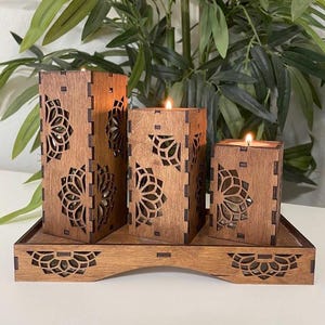 May include: A set of three wooden candle holders with intricate laser-cut designs. The holders are arranged on a matching wooden tray with a curved base.