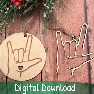 Puede incluir: Dos adornos de madera con forma de manos en el signo de la lengua de señas americana "Te amo". Los adornos están sobre un fondo de madera. Un adorno es redondo y el otro es una forma de mano más tradicional.