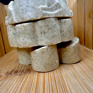 Puede incluir: Una pila de jabones hechos a mano con un color beige claro y una textura moteada. Los jabones están apilados sobre una bandeja de bambú, con un fondo de madera. Los jabones tienen una forma redondeada única, lo que sugiere un producto natural y orgánico.