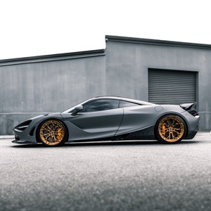 May include: A sleek, gray sports car with gold wheels parked in front of a gray building. The car has a low profile and a long hood.