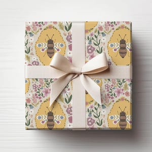 May include: A wrapped gift with a cream-colored ribbon and bow. The wrapping paper features a repeating pattern of bees in yellow ovals, surrounded by flowers in pink, purple, and green on a light beige background.